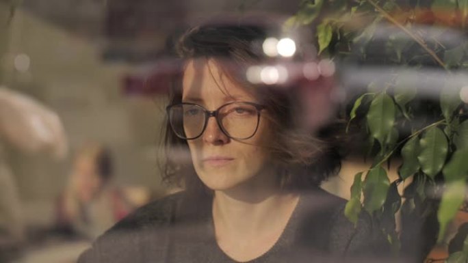 一个戴着眼镜的年轻女子在咖啡或修复剂中等待某人，以沉思，沉思的面部表情凝视着远方。汽车和人们在电影般