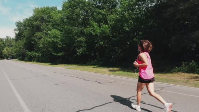 年轻迷人的女跑步者。穿着粉色衬衫的美丽上进心女孩正在城市公园跑步。带耳机的运动女性在公园训练