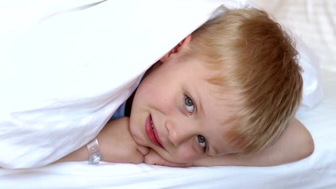 一个可爱的金发男孩在白色毯子下的床上，头上盖着毯子