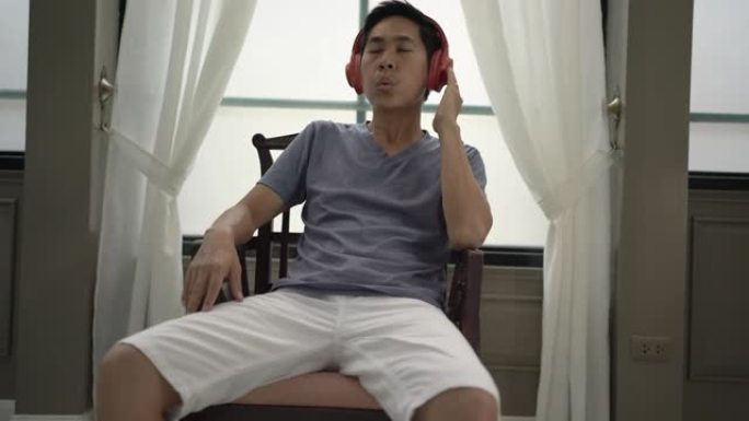 亚洲男子在椅子上放松，用白色窗帘在窗户旁听音乐。手持镜头，快乐男人概念