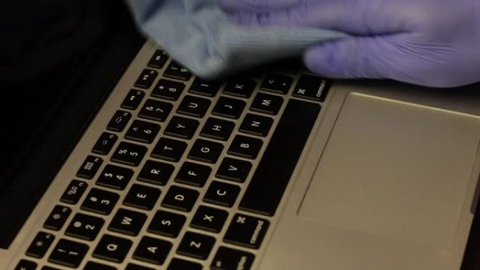 一个戴着橡胶手套的男人正在清洁他的笔记本电脑。将喷雾瓶中的清洁剂涂在纸巾上，然后擦拭笔记本电脑键盘。