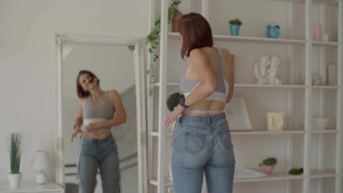 年轻女子在卧室穿着旧牛仔裤时欣赏减肥的结果。微笑的女性用厘米胶带在镜面反射下测量她的腰部。减轻重量
