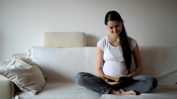 年轻的孕妇坐在家里的白色沙发上看书