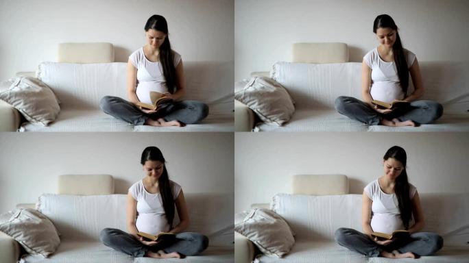 年轻的孕妇坐在家里的白色沙发上看书