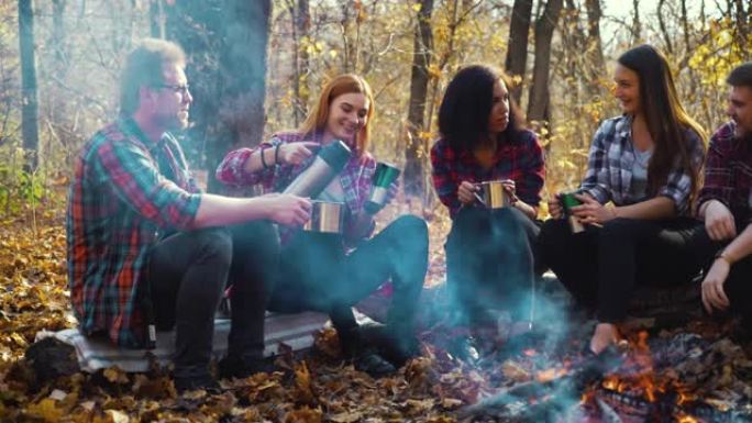在秋天的森林中，年轻的游客坐在火炉旁坐着保温杯