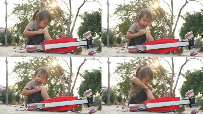 可爱的亚洲小女孩在夏季公园从踏板车上摔下来后坐在地上。幼儿健康户外运动。