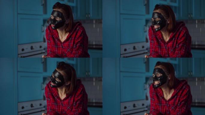 有趣的30多岁女人脸上戴着黑色化妆品面膜在家做鬼脸。女人在蓝色厨房的面具上做鬼脸。
