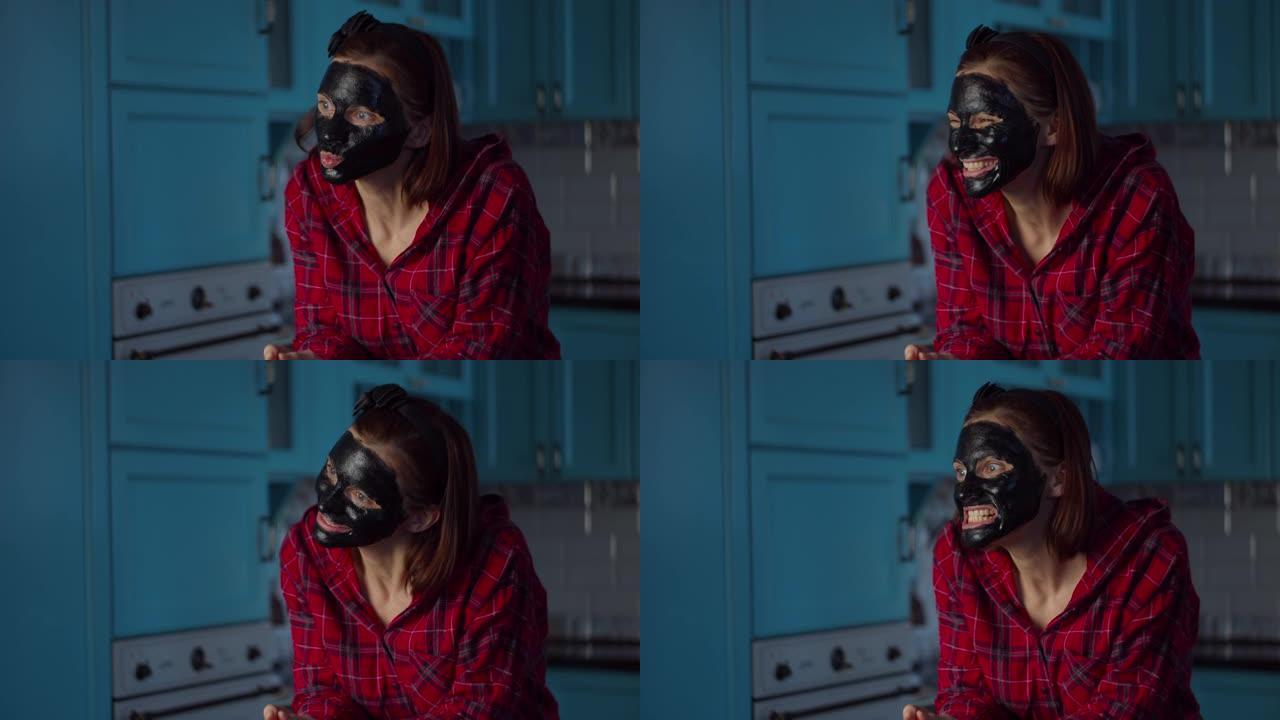 有趣的30多岁女人脸上戴着黑色化妆品面膜在家做鬼脸。女人在蓝色厨房的面具上做鬼脸。