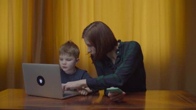 金发碧眼的学龄前男孩在笔记本电脑上打字，母亲拿着手机坐在家里的桌子上。孩子在网上学习，妈妈帮他。