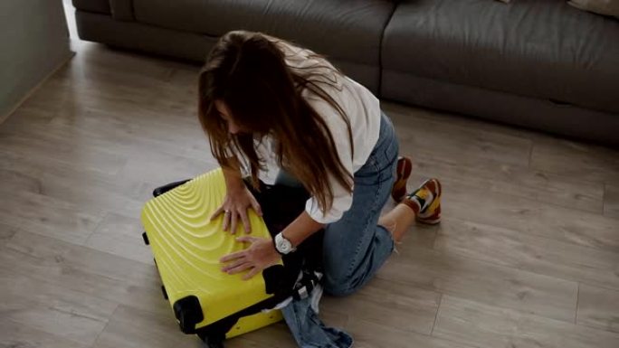 为旅行准备包。年轻女子打包行李，绝望地试图关闭装满的行李箱，并在其上拉上拉链，同时坐在客厅的地板上