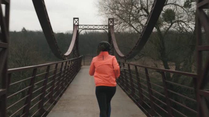 戴着耳机的女人正在公园里过桥。穿着橙色夹克和黑色紧身衣的年轻女运动员在户外过河的桥上奔跑。运动与健康