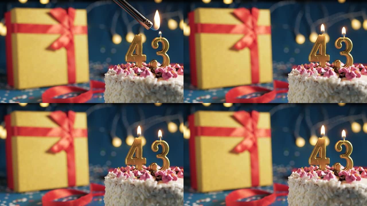 白色生日蛋糕编号43点灯燃烧的金色蜡烛，带灯的蓝色背景和用红丝带绑起来的礼物黄色盒子。特写