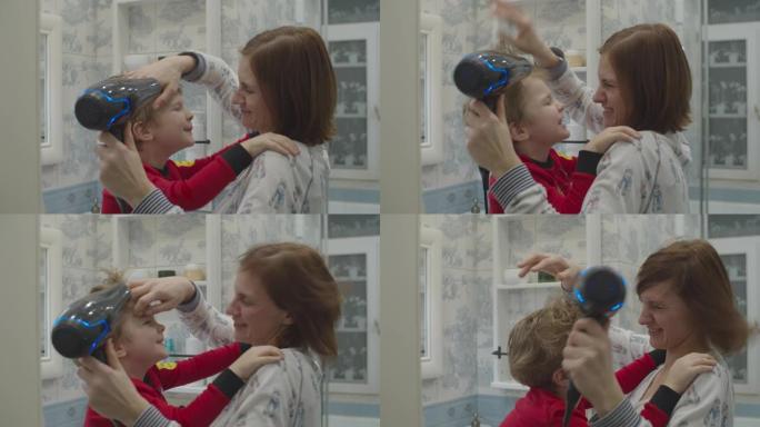 年轻的母亲用吹风机吹干儿子的头发。穿着红色睡衣的孩子在吹干头发时拥抱母亲。有趣的家庭沐浴程序。母子俩