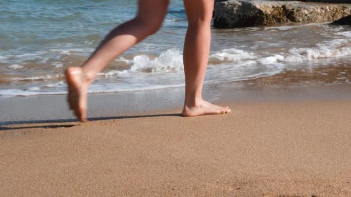 苗条女人的腿在黄沙上沿着大海行走。沙滩上晒黑的腿。年轻苗条的女孩沿着大海行走，在沙滩上留下脚印。