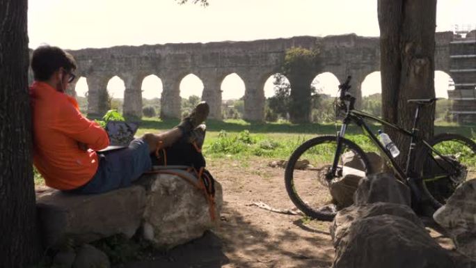 骑自行车的人坐在摄像机上的树下的一块岩石上，他的笔记本电脑在古罗马渡槽前。罗马公园里穿着橙色运动服的