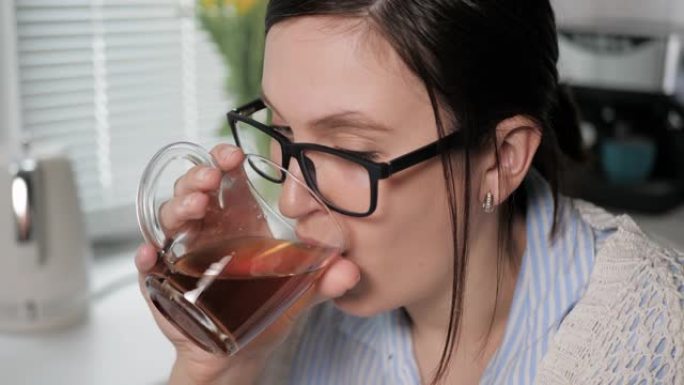 女孩喝绿茶或红茶。厨房里戴着眼镜的迷人年轻女子的特写镜头，慢慢地从透明杯子里喝棕色饮料，全神贯注地看