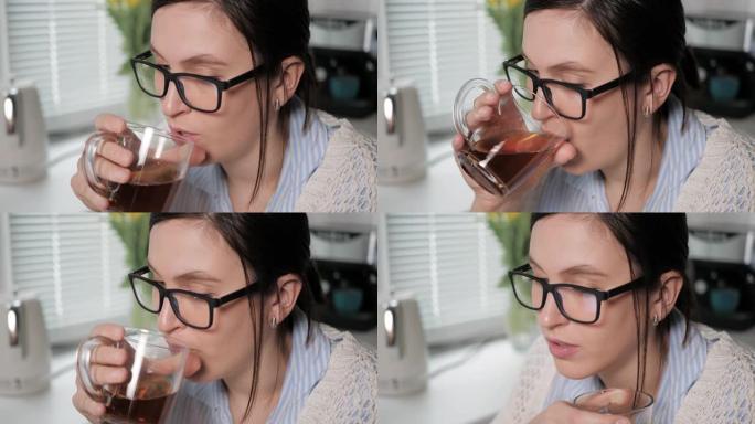 女孩喝绿茶或红茶。厨房里戴着眼镜的迷人年轻女子的特写镜头，慢慢地从透明杯子里喝棕色饮料，全神贯注地看
