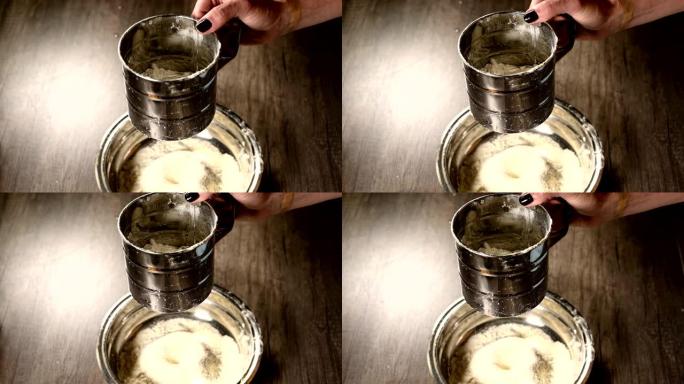 在家庭厨房的金属碗中，用筛子杯子用女性手筛面粉的特写镜头。烹饪家庭烘焙