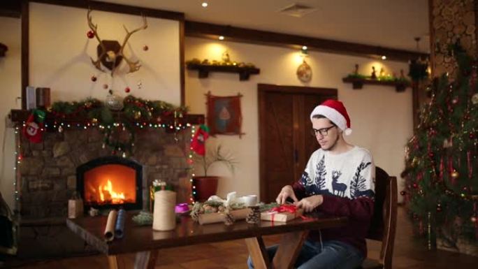 大胡子的男人坐在壁炉附近的新年礼物上鞠躬。戴着圣诞帽的家伙用纸包装礼物盒子，增加了冷杉树枝、圆锥体、