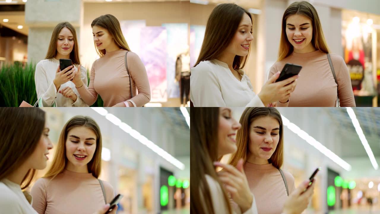 两个快乐的女性朋友带着购物袋浏览手机上的照片，有说有笑地站在商场走廊里，放大平移镜头