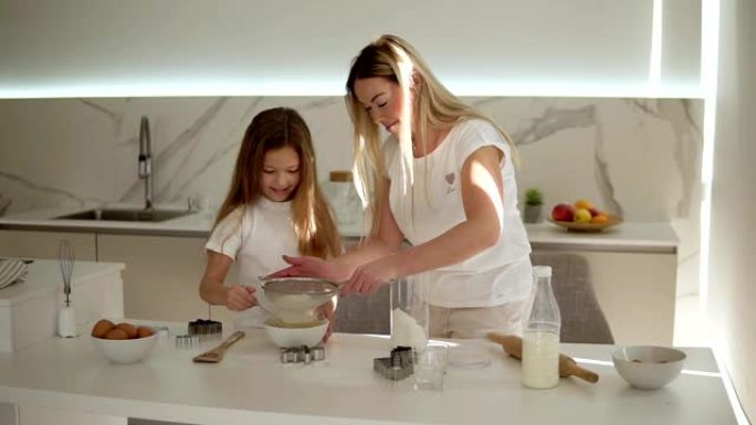 母亲在现代舒适的白色厨房里帮助和教她可爱的女儿做蛋糕或煎饼。母亲通过筛子和女孩混合配料加入面粉。幸福
