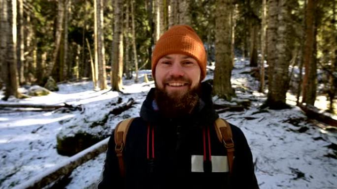 一个快乐的大胡子笑着的年轻人的肖像，他的背包站在针叶树的冬季森林中