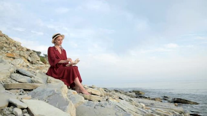 穿着夏季红色连衣裙和草帽的迷人微笑的高加索女孩的肖像，怀里抱着一本书，坐在海边的石头上，翻阅书页
