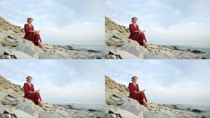 穿着夏季红色连衣裙和草帽的迷人微笑的高加索女孩的肖像，怀里抱着一本书，坐在海边的石头上，翻阅书页