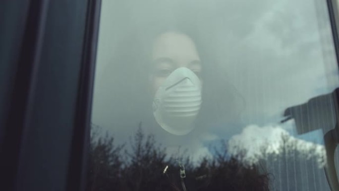沉思的年轻女子戴着口罩，在看着窗外时保护自己免受新型冠状病毒肺炎。一只飞行中的鸟被反射在窗户玻璃上