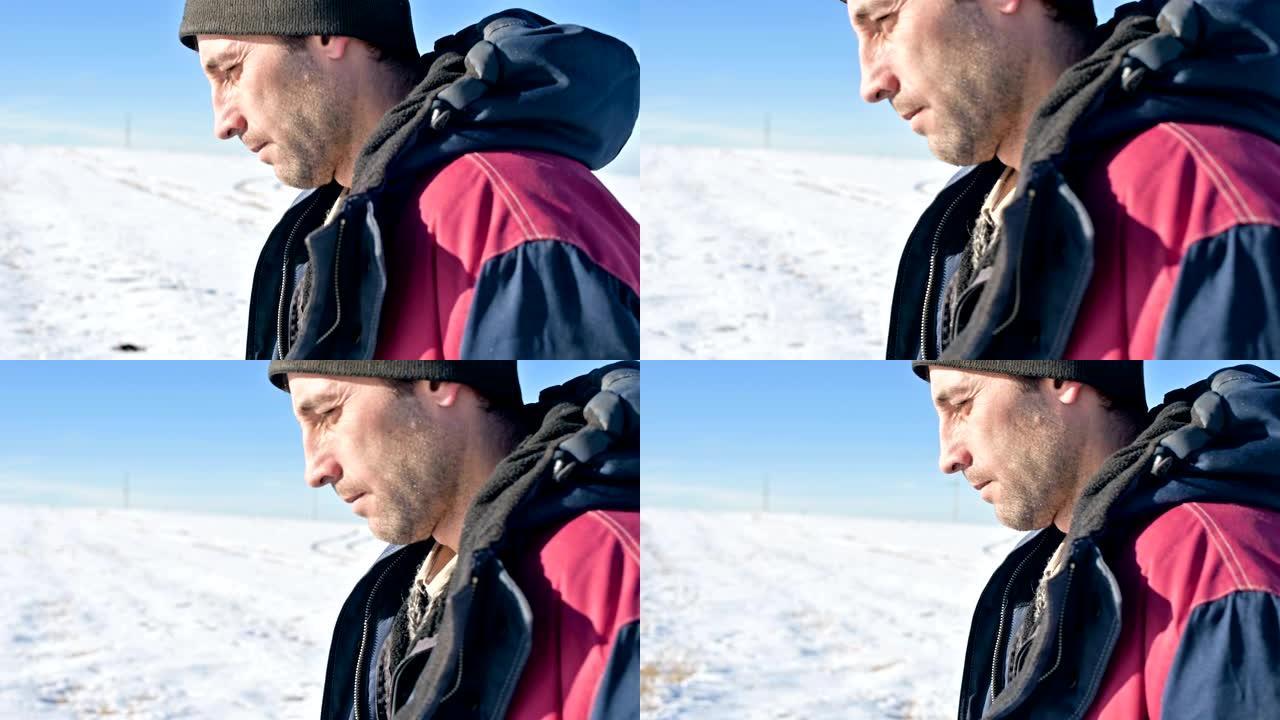 在一个阳光明媚的冬日里，一个戴着帽子和保暖外套的沉思男子的特写肖像，在白雪覆盖的户外田野中。若有所思