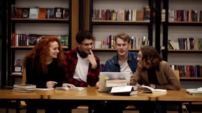 一群年轻的学生在图书馆一起学习，使用笔记本电脑讲解课程。积极讨论某事，笑着打手势，看着镜头。坐在木桌