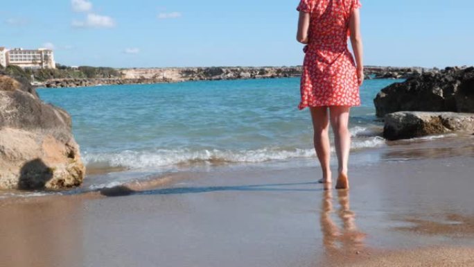 年轻的高加索女孩在海边下水。穿着红色短裙的黑发女孩走在海边。迷人的女人走过海边。穿着红色连衣裙的年轻