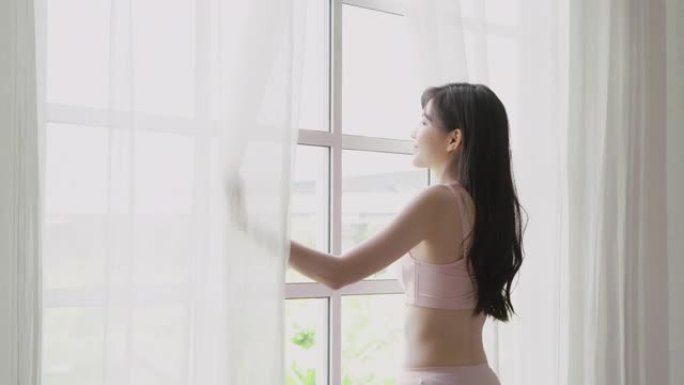 美丽的年轻亚洲女子醒来，早上带着打开的窗帘走到窗前，穿着舒适、性感的内衣女孩微笑着放松，在假期里以快