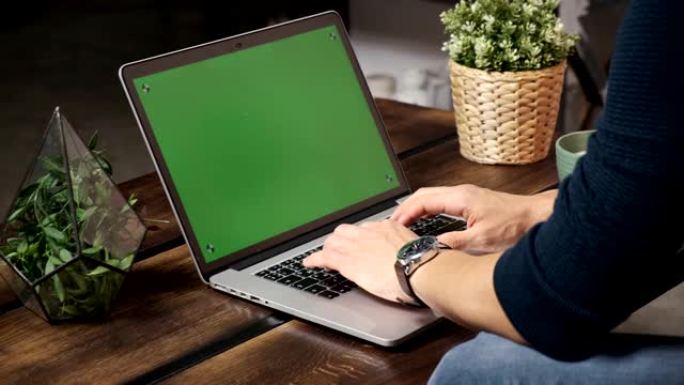 桌子上的人正在绿色的笔记本电脑屏幕上浏览互联网。在光线充足、舒适的公寓里。一名男子在阁楼办公室工作