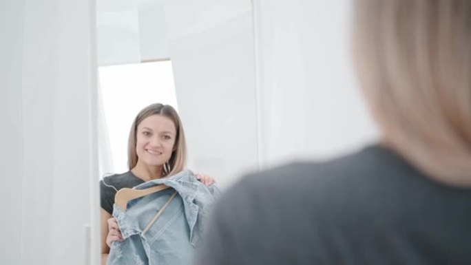 白色房间里一位年轻迷人的千禧一代女性试穿并选择衣服。照镜子，比较服装。房子的衣柜间或店内的试衣间。