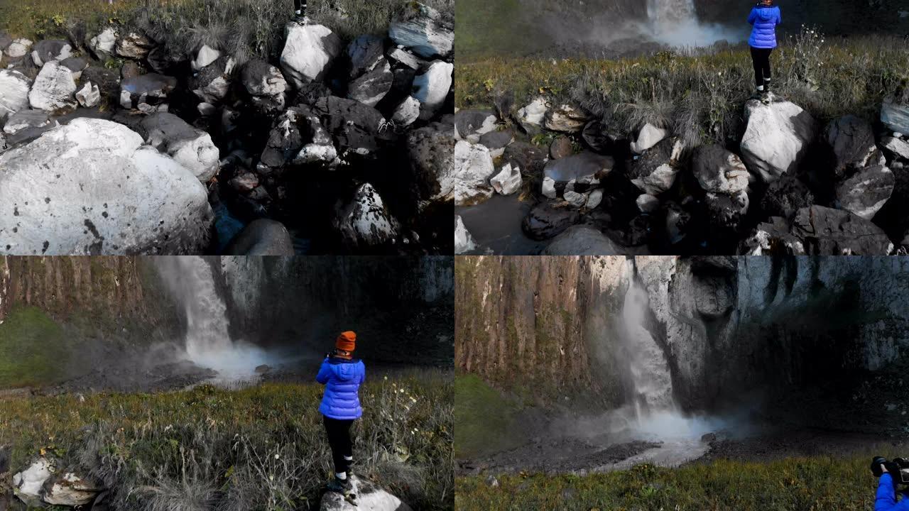 一个女孩是专业的风景摄影师，站在河边的石头上，拍摄大型史诗般的瀑布。鸟瞰图。北高加索