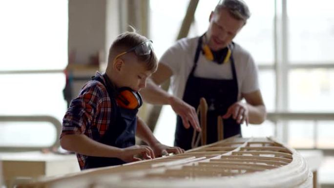 小男孩在木工车间的父亲帮助下建造大型木船模型的侧视图