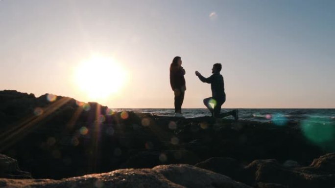 害羞的有魅力的男人站在膝盖上，在日落时向女友求婚。幸福的年轻夫妇在日落时在海边接吻和拥抱。求婚男人的