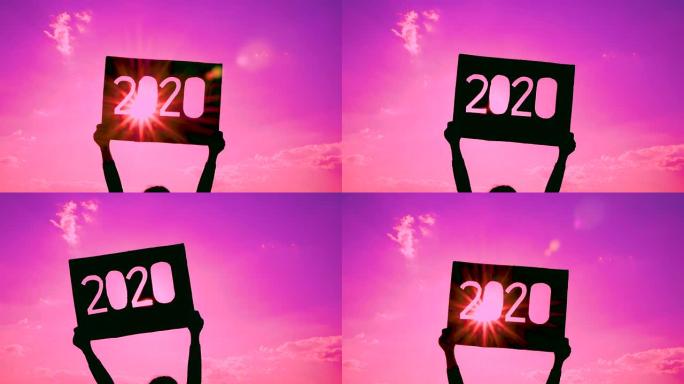 手握回收纸板的剪影，上面有粉红色的天空背景和阳光照耀着文本的2020文本。新年和庆祝活动的概念。