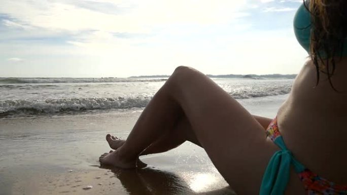 双腿纤细的年轻女子坐在海边附近，在阳光明媚的日子晒日光浴。晒黑的女孩在美丽的海滩上躺在潮湿的沙滩上。