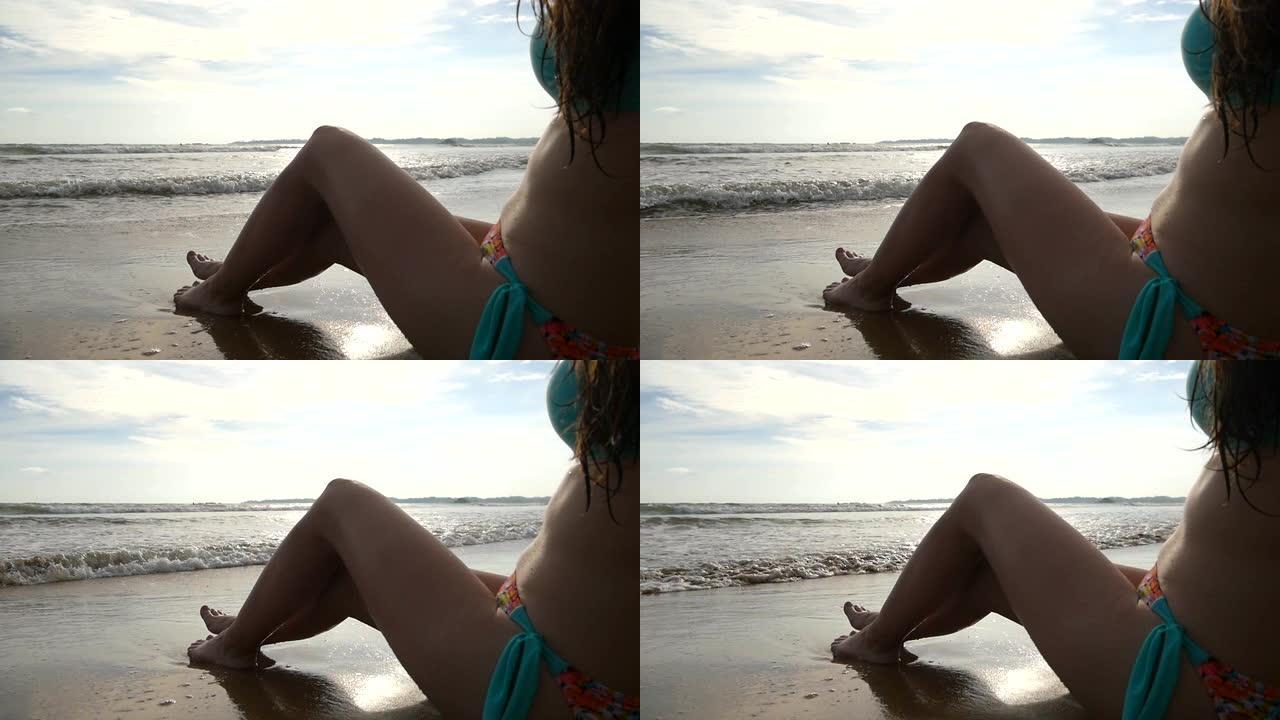 双腿纤细的年轻女子坐在海边附近，在阳光明媚的日子晒日光浴。晒黑的女孩在美丽的海滩上躺在潮湿的沙滩上。