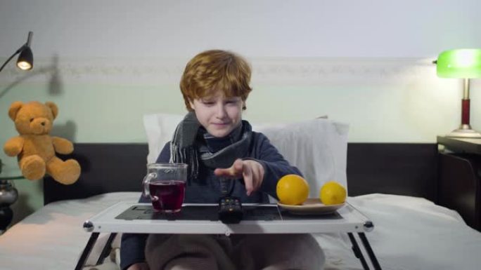 咳嗽的白人小男孩拿遥控器和切换电视频道的肖像。红发可爱的孩子坐在托盘前的床上，端着茶和橘子。疾病，健