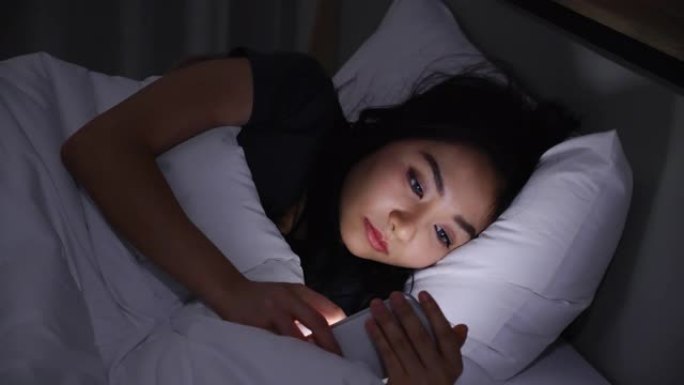 亚洲年轻女子躺在床上，晚上在卧室里关掉灯玩智能手机。在手机上阅读社交。在弱光下使用手机对眼睛有健康影