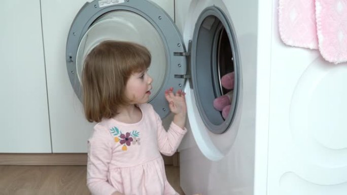 微笑的3岁小女孩的肖像坐在地板上，放在洗衣机的毛绒玩具中进行洗涤。儿童洗涤玩具