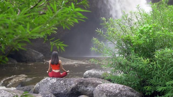 后视亚洲女性在瀑布练习或做瑜伽，莲花在冥想会上摆姿势。美丽的风景，自然的背景，泰国。