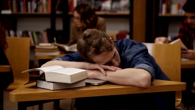 大学图书馆或班级里的年轻欧洲学生睡在桌子上，上面堆着书。背景上的同学和书架
