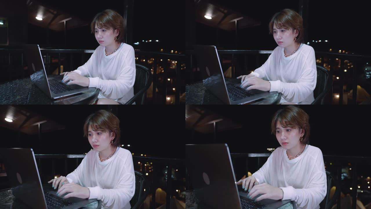 年轻的亚洲妇女坐在阳台上，晚上在笔记本电脑上工作