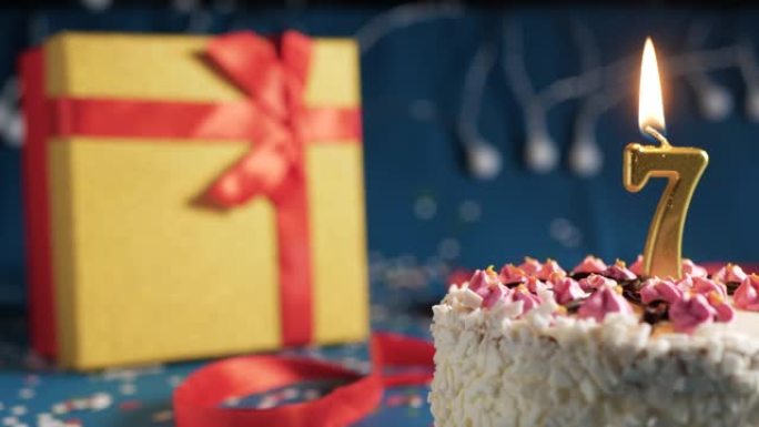 白色生日蛋糕7号点火器燃烧的金色蜡烛，蓝色背景带灯和用红丝带捆绑的礼物黄色盒子。特写