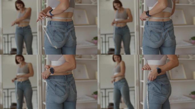 年轻女子在卧室穿着旧牛仔裤时欣赏减肥的结果。微笑的女性用厘米胶带在镜面反射下测量她的腰部。减轻重量