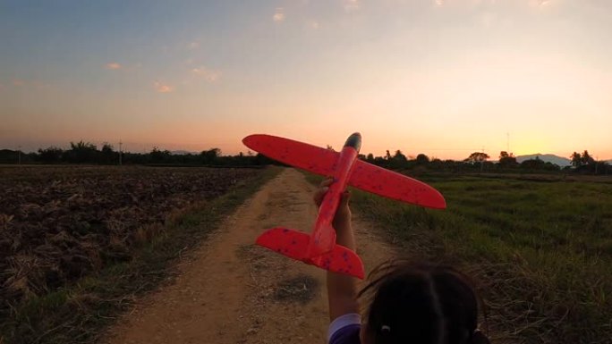 一个快乐的小女孩的慢动作在日落背景下与玩具飞机一起奔跑。幸福家庭和童年梦想的概念。